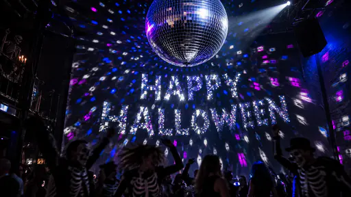 A large silver mirror ball hanging in a dark dance hall scatters blue, magenta and silver light patches over a wall that read HAPPY above HALLOWEEN, with dancers in skeleton costumes and face paint in silhouette at the bottom and a skeleton figure at the upper left.