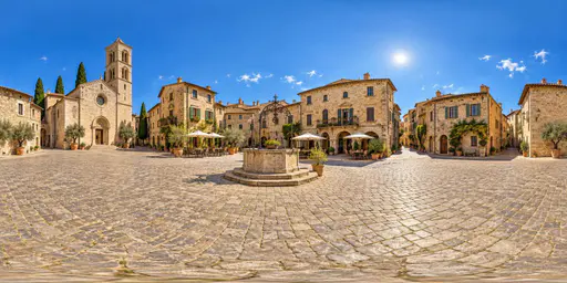 A full 360 degree equirectangular panorama from the centre of a historic stone town square with cobbled paving, a well, old townhouses with shuttered windows, a church tower and cafe tables with umbrellas.