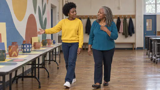 A young woman with a short natural afro in a yellow sweatshirt points with an open hand to a long table of plain painted pots and paper shapes while a full-figured woman with silver locs in a teal blouse nods with a hand at her pendant in soft light.