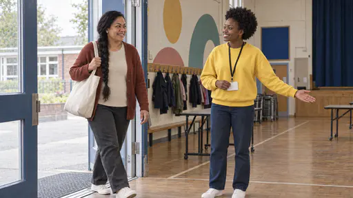 A young woman with a short natural afro in a yellow sweatshirt holds out a welcoming hand and a blank lanyard card to a woman with a long black braid and a tote bag stepping in at the open door of a school hall in bright light.