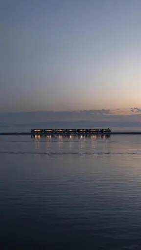 Seven amber-lit carriage groups cross an embankment near the middle of a tall dawn landscape; rippled dark water reflects the lights.