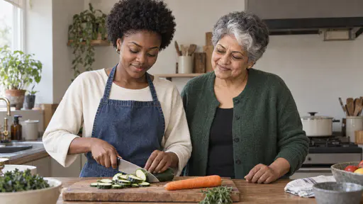 A young woman with a short natural afro in a denim apron carefully slices a carrot on a shared wooden board beside a South Asian woman with short silver-grey hair and a white streak who watches with a pleased nod at a kitchen counter in warm midday light.