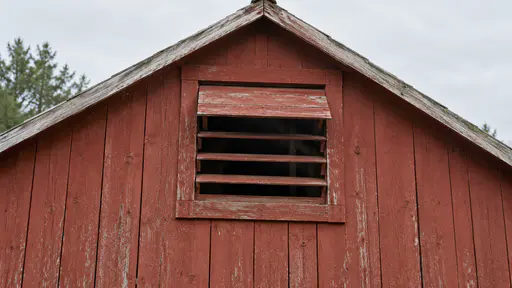 The loft ventilation opening beneath the roof of a red barn shows its slatted cover partly open in clear daylight.