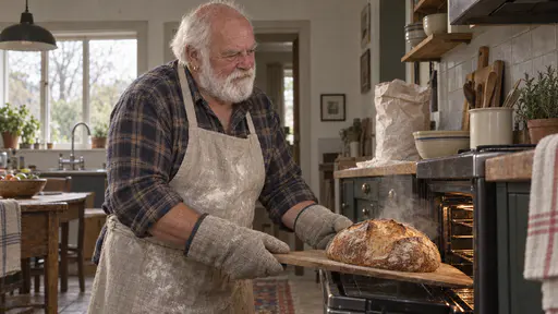 A broad white-bearded man with a bald crown and a gold hoop earring in a floury apron and thick oven gloves draws a golden loaf of bread out of an open oven on a wooden peel with steam rising and a bag of flour and a bowl on the counter behind him in late-afternoon light.