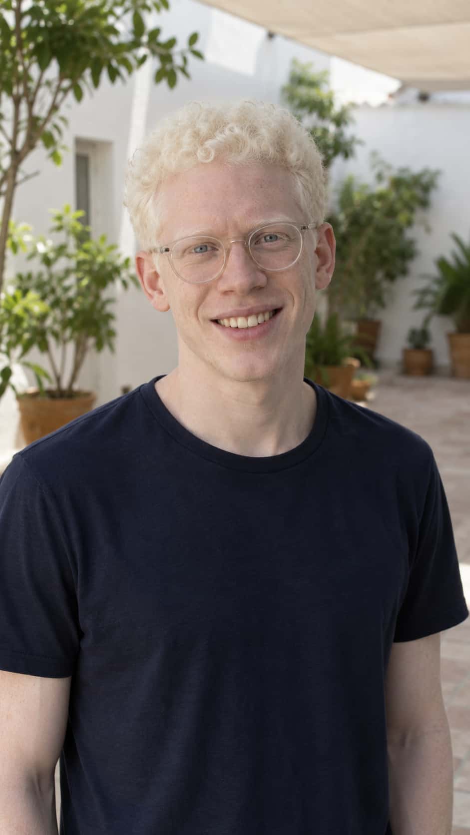 A slim smiling man in his late twenties with very pale ivory skin, short white-blond curly hair, light blue-grey eyes and plain round clear glasses, in a navy t-shirt in the open shade of a whitewashed courtyard with potted plants.