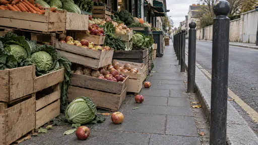 A tall heap of plain crates of vegetables and fruit spills from a market stall onto the pavement with a cabbage and apples rolled into the walkway and bollards beyond narrowing the walkway to one tight line in bright market light.