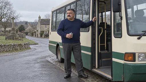 A heavyset bus driver with a wide moustache in a navy sweater stands beside the open front door of a small cream and green rural bus holding a flask on a quiet village green in cool early light.
