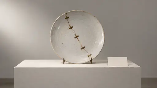 A plain ceramic dinner plate upright on a museum stand has one long crack closed by a row of small flat metal staple mends, with a blank label card beside it.