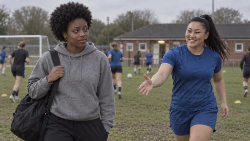 A slim young woman with a short natural afro and a small gold nose stud in a grey hoodie stands at the edge of a worn grass football pitch with a sports bag as a powerfully built woman in a blue training top with a high ponytail jogs over with an open hand, players warming up and a brick clubhouse behind them.