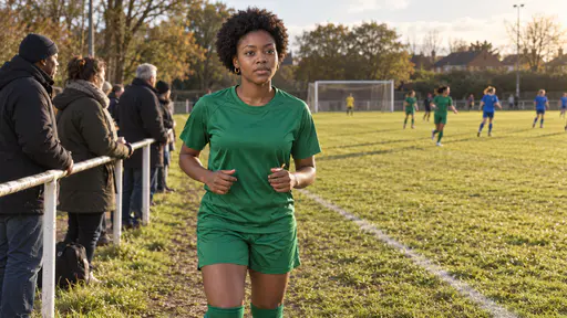 A slim young woman with a short natural afro in a plain green match shirt and shorts jogs on the spot on the touchline of a grass football pitch on match day, shaking out her hands, spectators in coats along the touchline behind her and a white goalpost beyond in late-afternoon sun.
