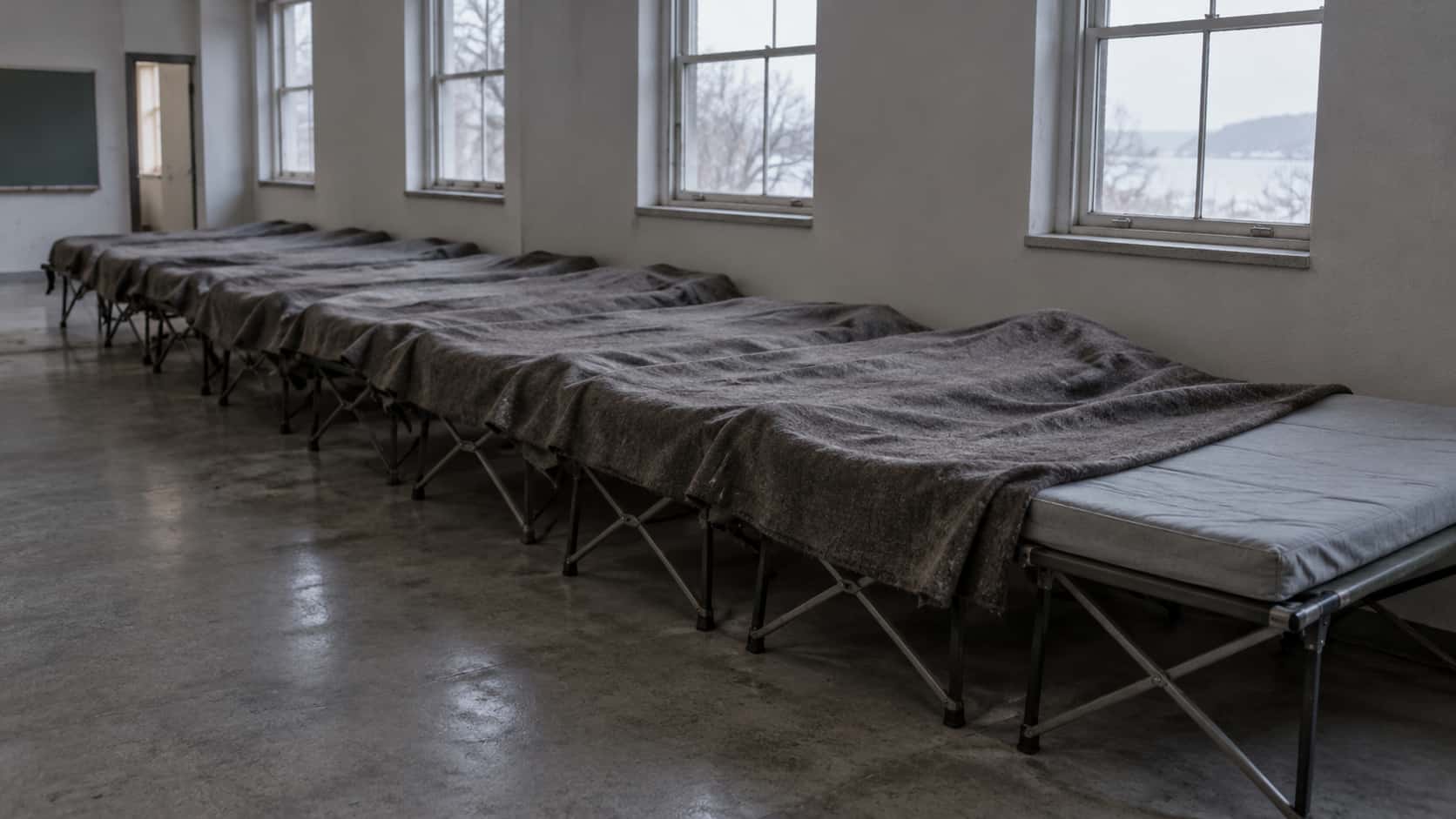 A long plain wool blanket is stretched across a row of eight narrow camp cots in a quiet hall, tucked over the first seven and just reaching the last cot with its bare grey mattress in soft grey dawn light.