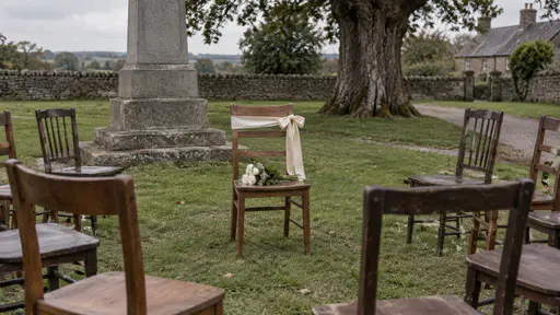 One plain wooden chair stands empty in a circle of chairs on a village green near a stone memorial with a blank cream cloth ribbon tied around its backrest and a few flower stems on its seat in flat overcast light.