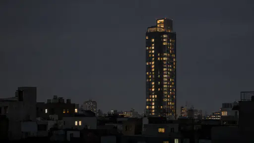 Low dark rooftops fill the lower frame at night beneath one single tall high-rise rising far above them with lit windows up its face.