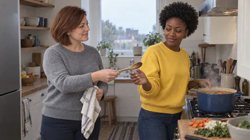 A white woman with a chin-length auburn bob in a heather-grey sweater hands a wooden spoon to a young woman with a short natural afro in a bright yellow sweatshirt who steps in to stir a pot of soup on the stove in a bright kitchen as steam rises.