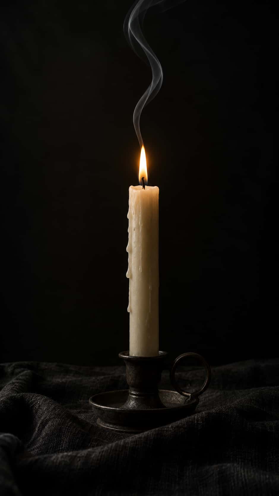 A cream taper candle with wax rings and a small steady flame, a thin ribbon of grey smoke curling up, dark iron holder, black linen, deep black background.