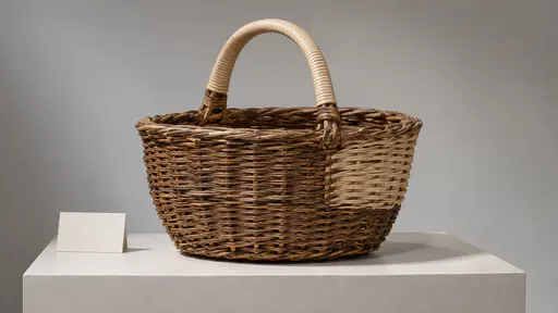 A wicker shopping basket on a museum plinth shows one side section rewoven in pale cane against the darker aged weave, with a blank label card beside it.