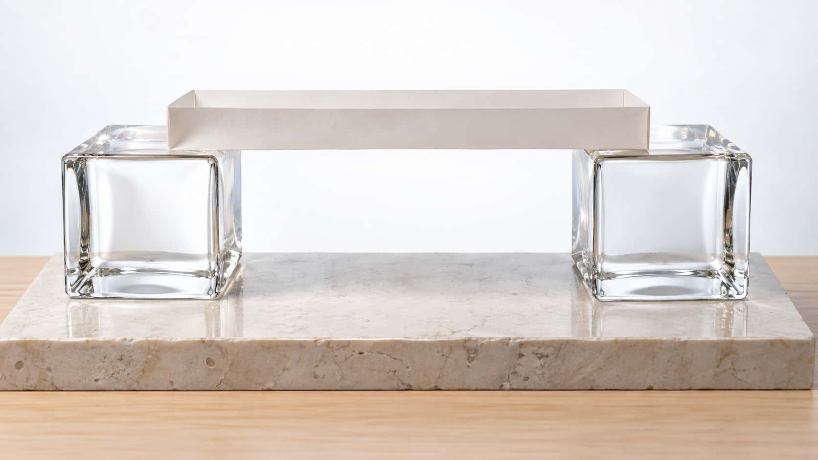 A folded white paper bridge deck with low side walls rests across the tops of two square clear glass block piers on a pale stone plinth with a pale wooden base, held up a hand's height above the plinth in bright backlight.