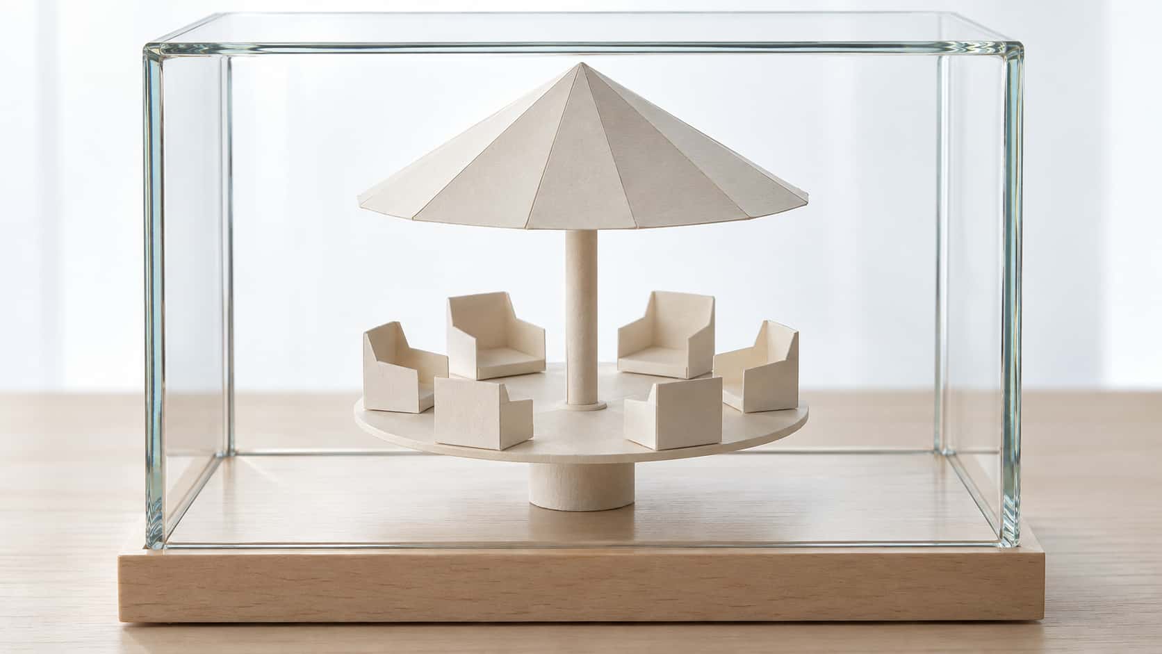 A flat round paper platform on a short central paper post carries six small folded paper seats around its edge under a paper canopy cone joined by a thin paper rod inside a clear rectangular glass display case on a pale wooden base.