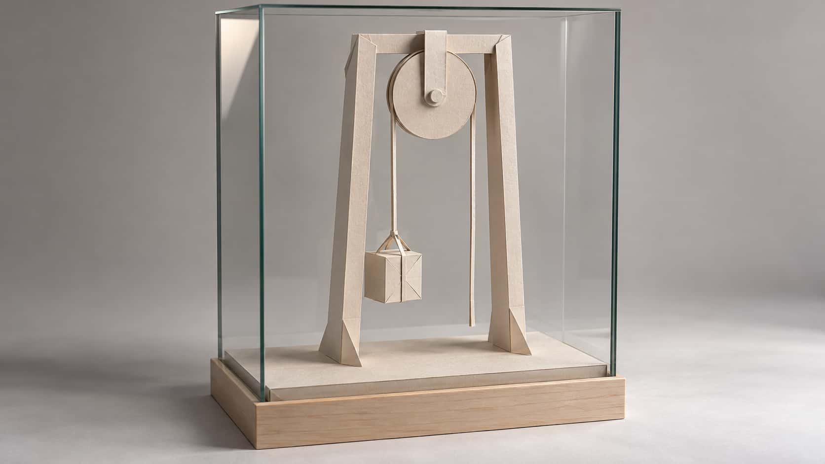 A large round paper pulley wheel on a bracket at the top of a tall paper frame carries a thin paper rope that hoists a small folded paper crate off a paper floor, the free end hanging straight down, inside a clear rectangular glass display case on a pale wooden base.