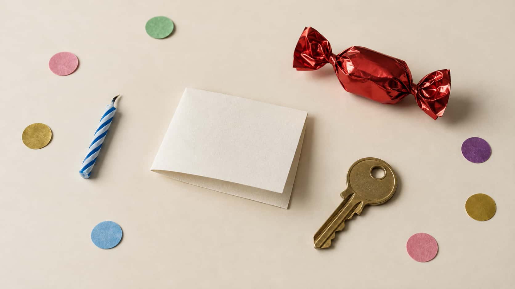 A flat-lay on cream paper of a few pieces of coloured confetti, a short striped birthday candle stub, a blank folded paper note, one sweet wrapped in red foil and a single brass door key with empty space around them.