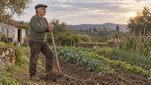 A broad-shouldered weathered man with a grey moustache in an olive-grey sweater and a flat cap leans on a long-handled wooden hoe with a dark soil-stained blade at the edge of a vegetable plot looking calmly across the rows in low golden evening light.