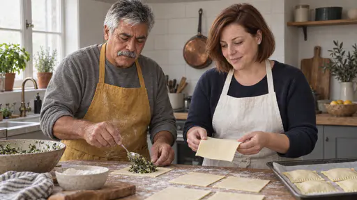 A man with a grey moustache in a mustard apron spoons green spinach filling onto the centre of a square of rolled pastry while a woman with an auburn bob in a white apron holds a second square flat ready to fold over it beside a lined baking tray.