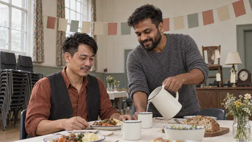 A man with swept hair in a rust shirt sits at a table with a plate while a broad man in a grey jumper leans in to serve him from a jug with a mended chair, a lamp and a clock displayed on a sideboard behind in diffuse hall light.