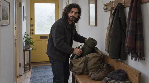 A bearded man in a charcoal jacket loads a rolled sleeping mat and a thermos into a large canvas holdall on an oak bench in a hallway beside a pair of boots and a hat in cool daylight through a frosted door panel.