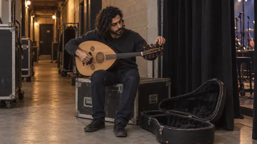 A slim man with thick wavy black hair and a full dark beard in a charcoal sweater sits on a flight case tuning a small acoustic oud with his head tilted to listen in a low amber backstage corridor beside a black curtain and an open instrument case at his feet.