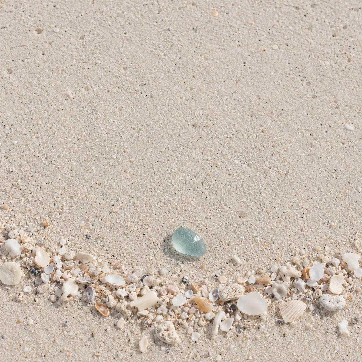 Pale beach sand seen from above with one thin curved tide line of fine shells and chalk fragments across the bottom fifth; a single piece of soft blue-green sea glass rests just above the arc on untouched sand, catching one small bright highlight, while the top of the frame stays clear.