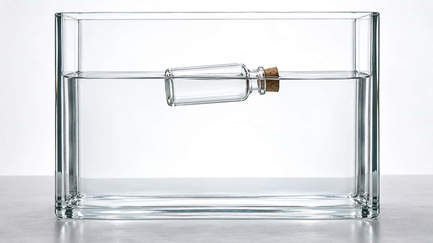 A small sealed empty glass vial with a cork floats on its side across the waterline of a wide clear glass tank of water in front of a bright diffused backdrop.