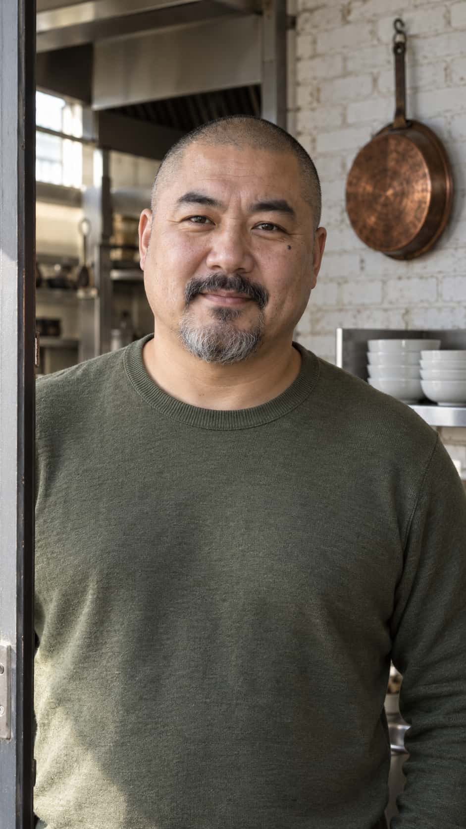 A man in his mid forties with a shaved head, salt-and-pepper goatee and a small mole under one eye, in an olive-green sweater in a kitchen doorway beside a whitewashed brick wall with a hanging copper pan and white bowls.