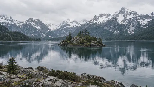 A small rocky island sits in a mountain lake below high snow-dusted peaks with their reflection faint in the still water.