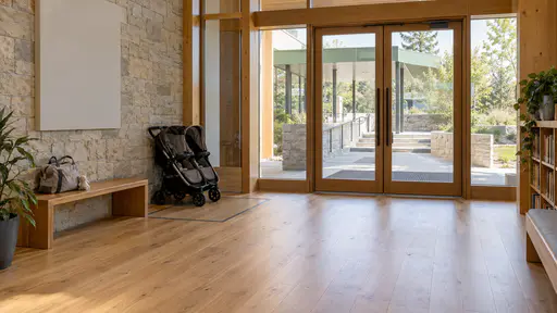 An entrance lobby has a parked double pushchair against the wall, a clear wheelchair space marked by a plain floor inlay and a low bench with a baby bag beside a glass door in soft diffuse light.