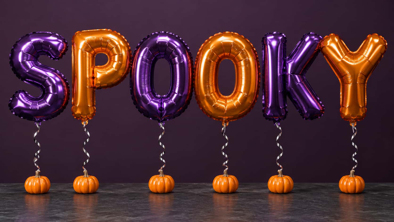 Giant foil balloons spelling SPOOKY in alternating purple and orange, each with a curled silver ribbon tied to a small orange pumpkin on a dark slate table against a plum backdrop.