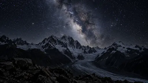 A dense star field and the Milky Way rise above jagged snow peaks with a faintly glowing glacier tongue and dark foreground rocks at night.