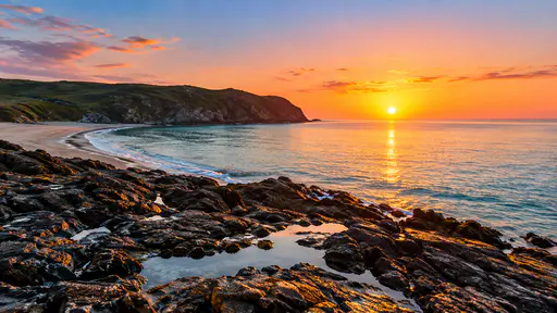 The sun just clears the horizon over a calm sea laying a golden glitter path past wet dark rocks, tide pools and a curved bay of pale sand.