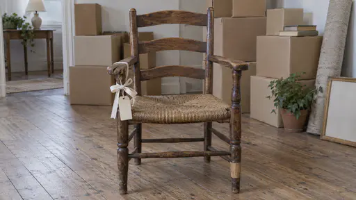 A worn oak armchair with a rush seat stands in a half-cleared room with stacked moving boxes behind it and a cream ribbon tied in a bow around one armrest with a small blank tag hanging from it in soft diffuse light.