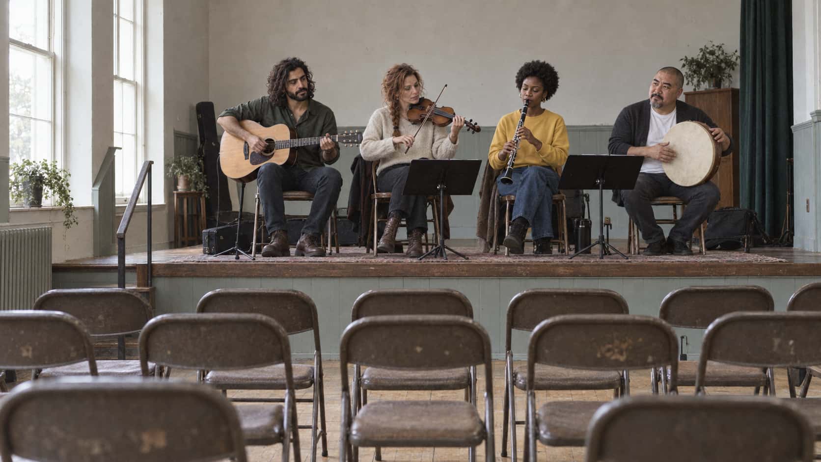 Four musicians with a guitar, a fiddle, a clarinet and a frame drum perform on a low stage in a community hall in bright side light from high windows with rows of empty folding chairs facing them.