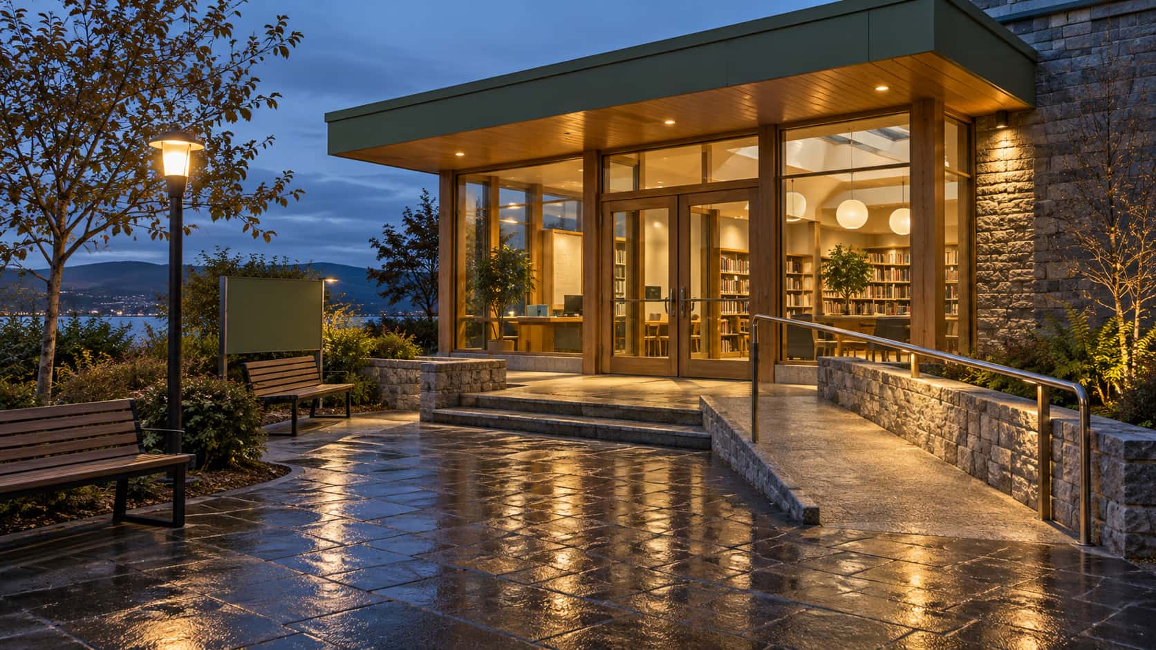 A small library entrance glows warm through its glass wall under a sage green canopy at blue dusk with the ramp handrail catching the light, a lamp on a post by the benches and a dark wet paved approach.