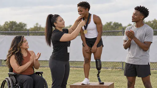 A powerfully built woman with a thick black high ponytail hangs a plain ribbon medal round the neck of a beaming lean runner with close-cropped hair and a running blade on a low podium while a woman in a manual wheelchair and a tall lean young man applaud beside it in warm afternoon light.