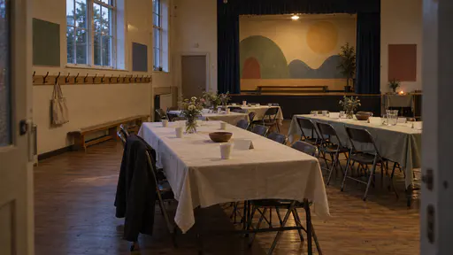 An empty small primary school hall after a welcome evening with long folding tables pushed together under plain cloths, blank name cards and cups left behind, one forgotten canvas bag on the coat pegs and a painted mural glowing under the last warm lamp.