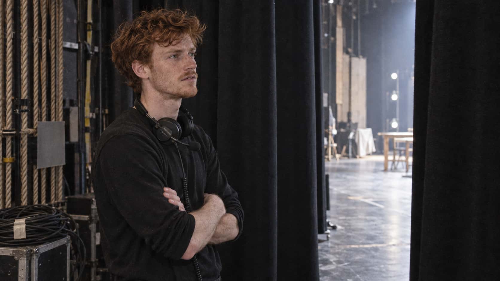 A freckled red-haired stagehand in black stands with arms crossed and a headset round his neck in the wings backstage beside black curtains, a fly rail and a coiled cable in cool light spilling from the stage.