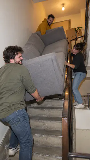 Three fictional adult friends in casual clothes laugh as they angle a grey two seat sofa up a narrow apartment stairwell past boxes on the landing in a compact camera flash photograph.