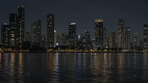 A continuous city skyline of dark towers with scattered lit windows stretches behind a wide dark river at night, weak reflections broken on the water.