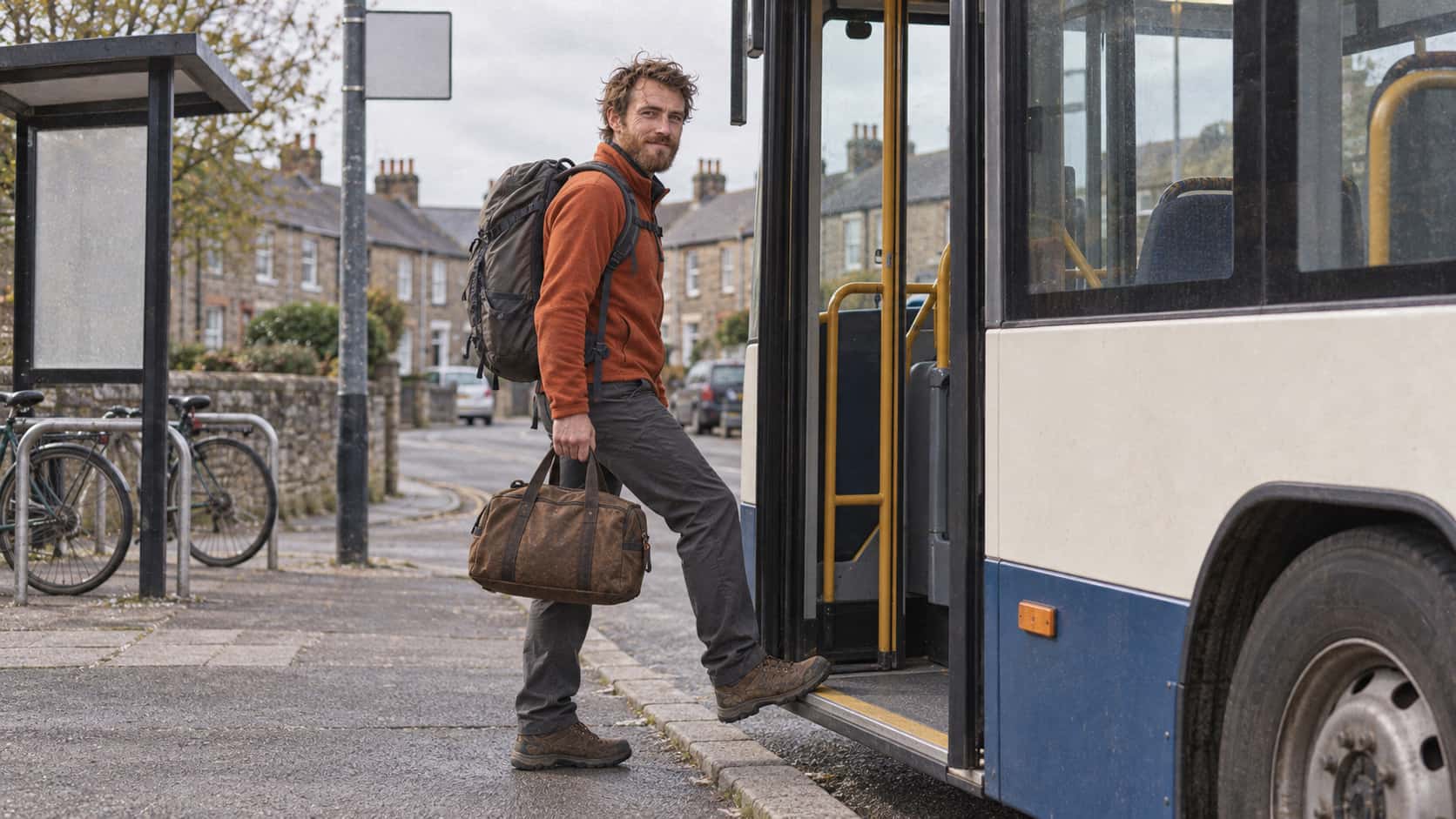 A lean man with an auburn beard in a rust fleece with a grey rucksack stands on the kerb at a small town bus stop beside a bus whose doors have just folded open with a small brown duffel bag held up ready to step on, a row of terraced houses behind in bright midday light.
