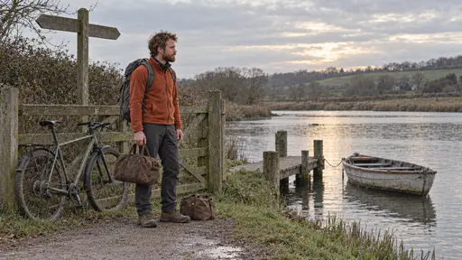 A lean man with an auburn beard in a rust fleece stands with his bicycle leaning against a gate and a small brown duffel bag at his feet at the end of a country lane where a quiet river with a small wooden jetty and a rowing boat meets a footpath with a blank signpost in soft evening light.