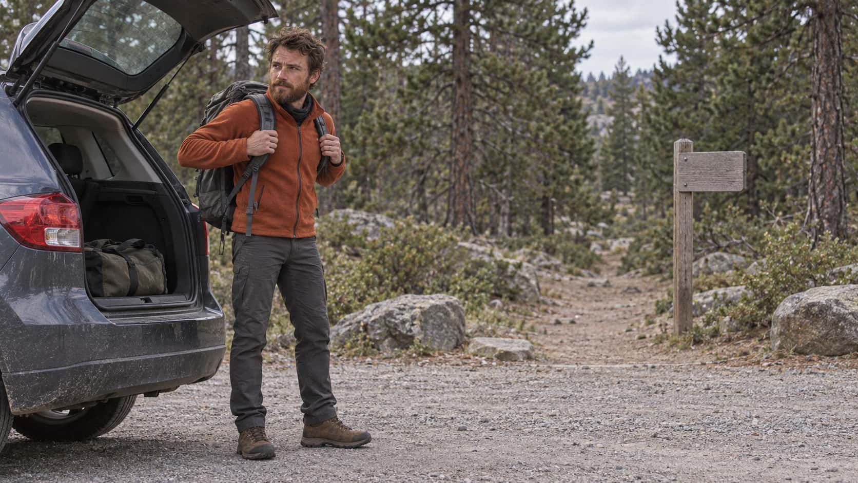 A lean man with an auburn beard in a rust fleece and walking boots settles a grey rucksack onto his shoulders beside the open boot of a small hatchback at a trailhead car park at the edge of a pine forest with a trail opening between the pines and a blank marker post in soft afternoon light.