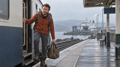 A lean man with an auburn beard and tousled brown hair in a rust fleece with a grey rucksack steps down from a stopped train carriage onto a quiet platform with a small brown duffel bag at his feet and a ferry terminal in a grey harbour beyond the platform in cool early morning light.