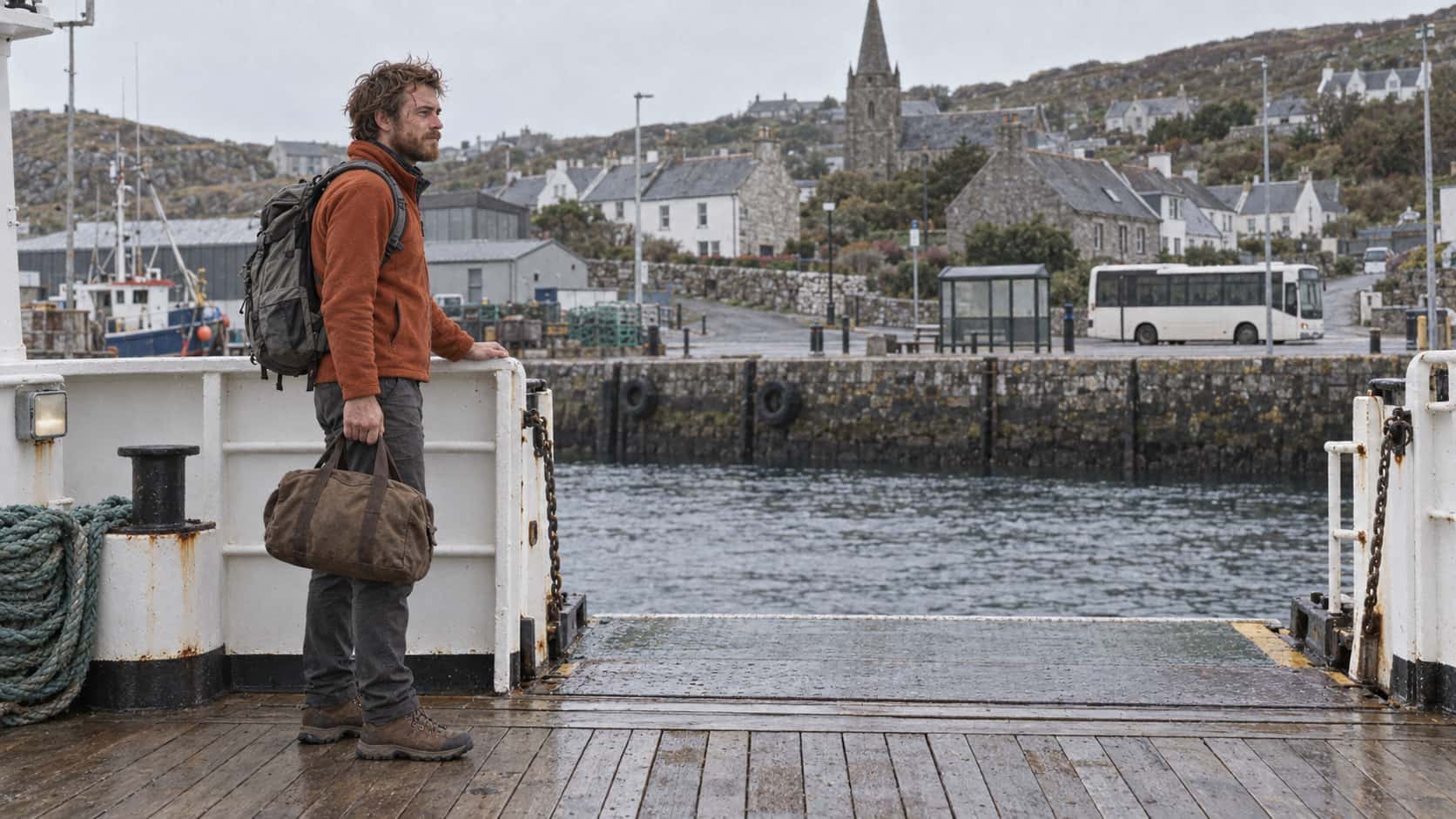 A lean man with an auburn beard in a rust fleece with a grey rucksack stands at the open gangway of a small ferry at a quay holding a small brown duffel bag in one hand and the rail in the other looking toward a harbour town and a bus waiting at the stop beyond in soft grey light.